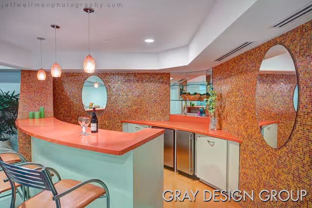 Interior view of a modern bar area with a coral-colored countertop, three hanging pendant lights, two bar stools with metal frames and tan seats, mosaic tile walls with circular mirrors, and a small refrigerator under the counter.
