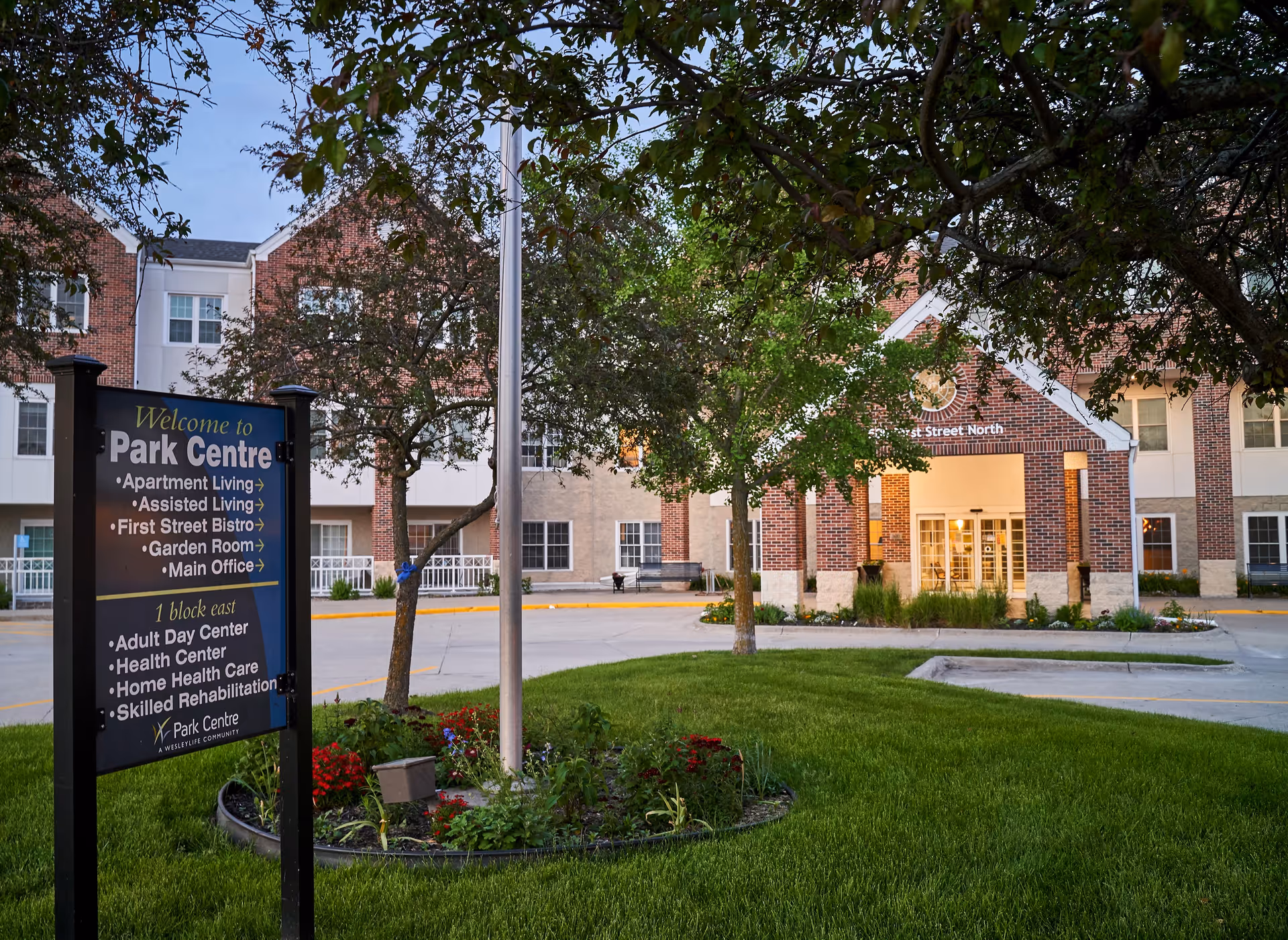 Front entrance of the Park Centre senior living building with a welcome sign, flagpole, trees, and landscaped lawn.