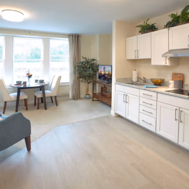 A bright and clean senior living space featuring a small dining area with a round table set for two near large windows, a kitchen area with white cabinets and granite countertops, and a living area with a TV on a wooden stand and a potted plant in the corner.