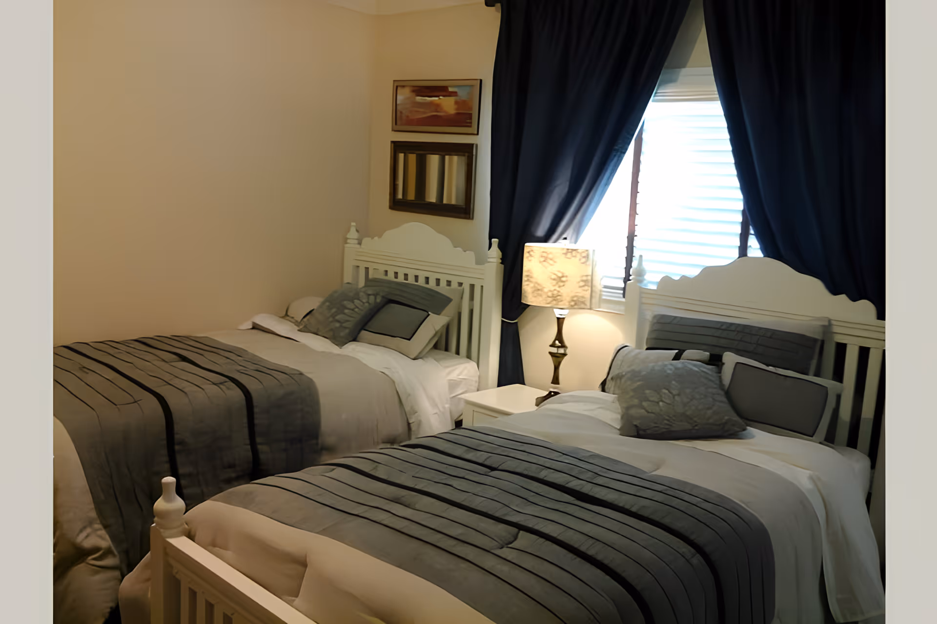 A bedroom with two white wooden beds, each made with gray and white bedding and multiple pillows. Between the beds is a white nightstand with a decorative lamp. A window with dark blue curtains is behind the nightstand, and two framed pictures hang on the wall above one of the beds.