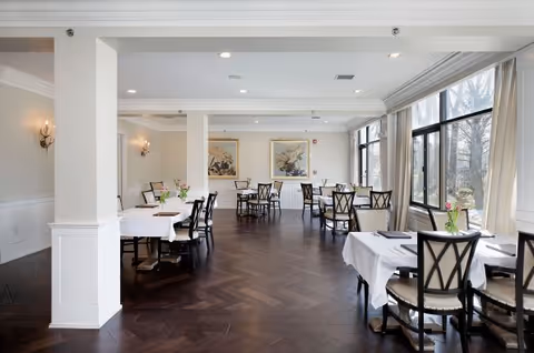 Spacious dining room with white-tablecloth tables, dark wooden chairs, large windows, and framed artwork on the walls.