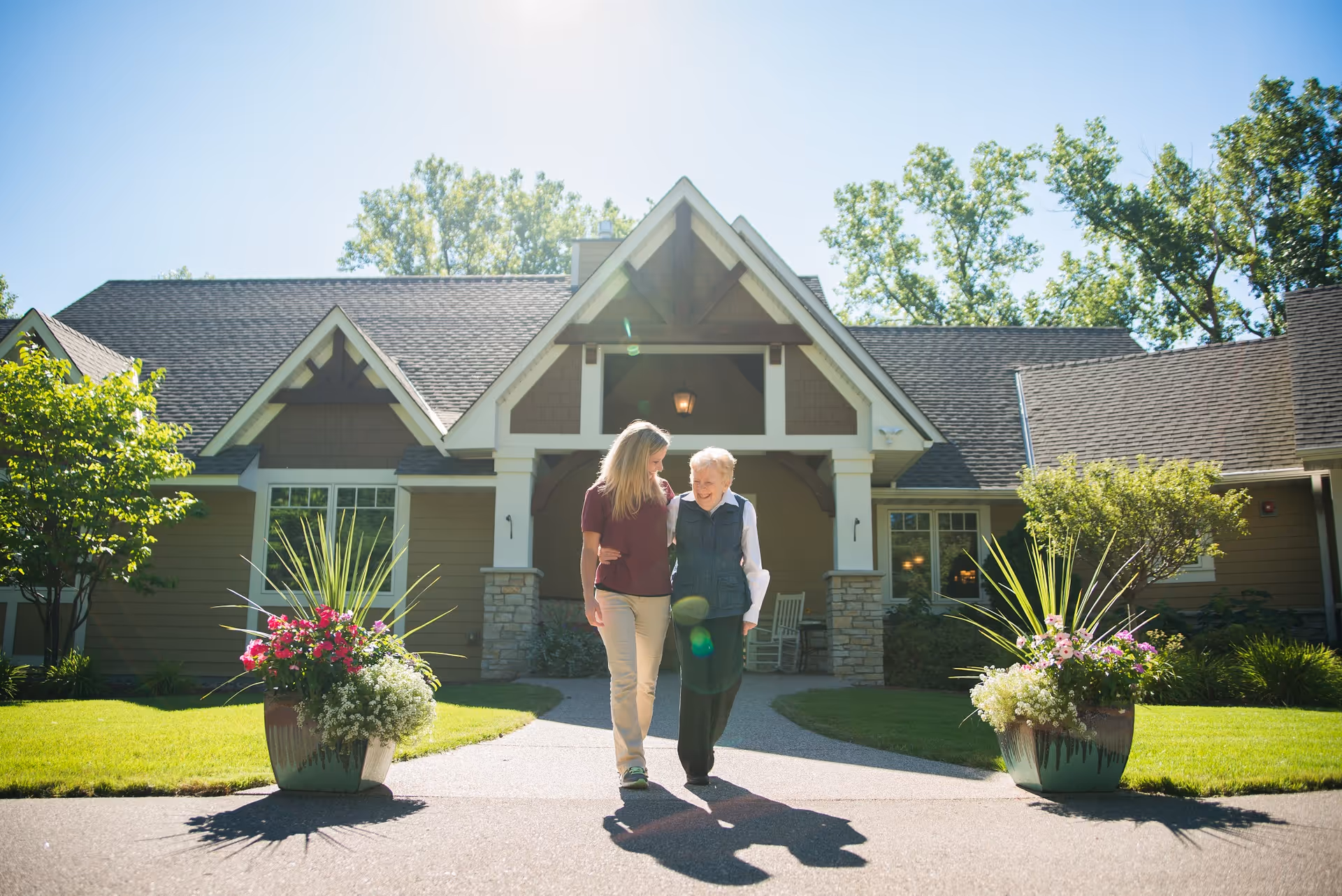 Two women, one elderly and one younger, walking arm in arm on a paved path in front of a large, well-maintained house with a peaked roof and stone accents. There are large flower pots with colorful flowers on either side of the path and green trees and grass surrounding the house.