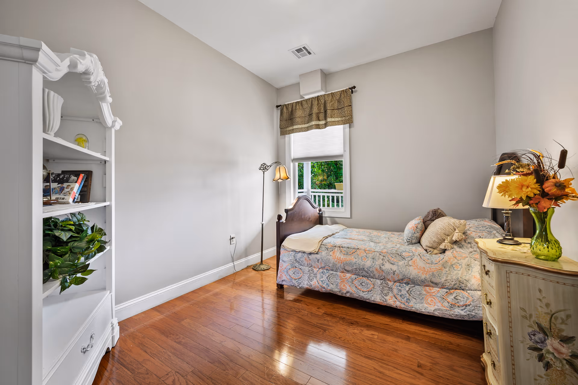 A small bedroom with a single bed covered in a patterned quilt and multiple pillows. There is a window with a valance and a white blind, a tall floor lamp beside the bed, a white bookshelf with plants and books on the left, and a small decorative dresser with a lamp and a vase of flowers on the right. The room has wooden flooring and light-colored walls.