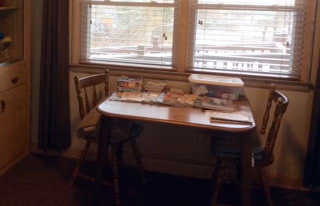A small wooden table with two wooden chairs placed in front of a window with blinds. The table is cluttered with various papers, magazines, and a plastic container. The room has wooden furniture and a curtain on the left side.