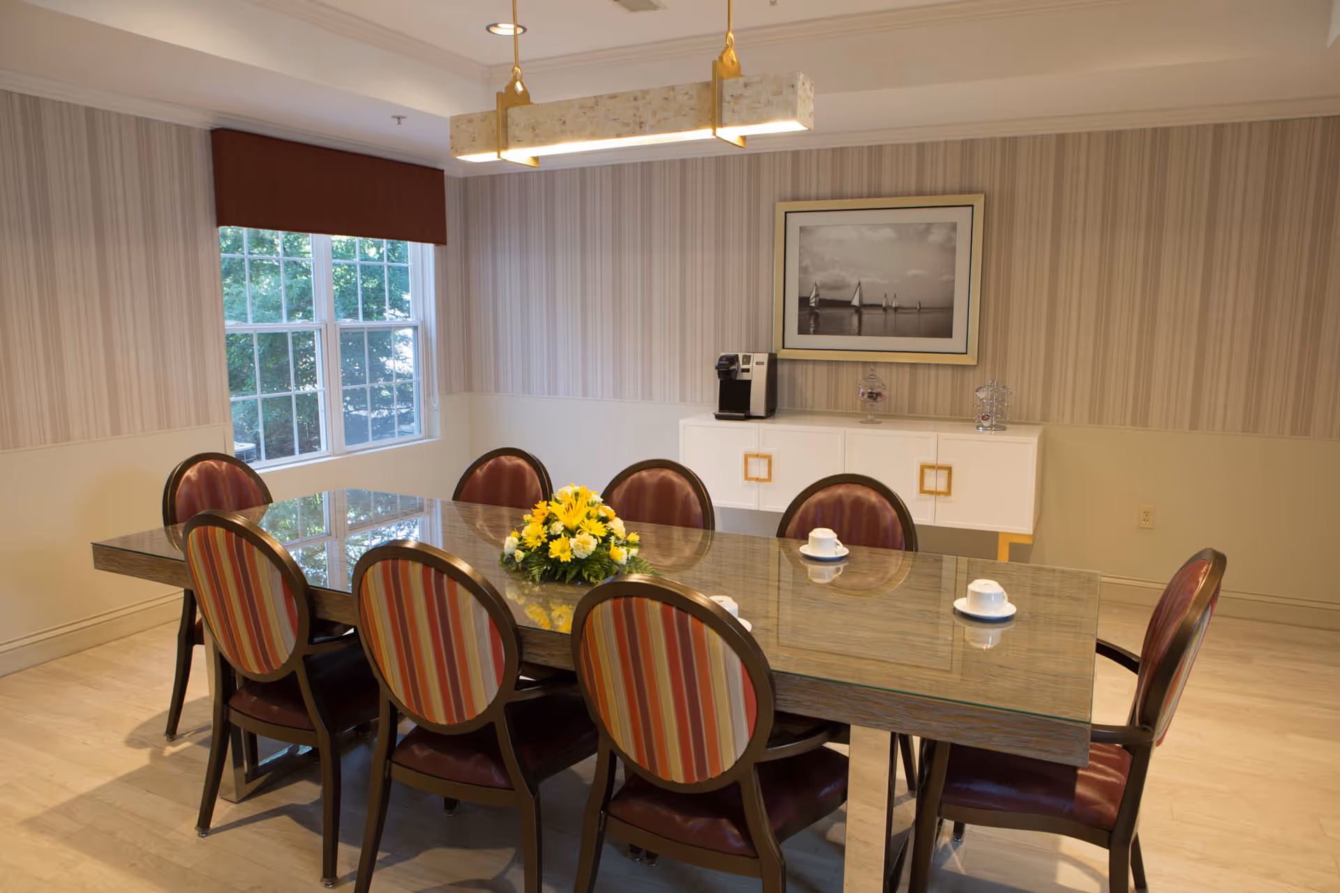 A dining room with a rectangular glass-top table surrounded by eight chairs with striped upholstery. A yellow flower arrangement is centered on the table. The room has a large window with a brown valance, striped wallpaper, and a white sideboard with a coffee machine and decorative items. A framed black and white photo of sailboats hangs above the sideboard. A modern rectangular ceiling light fixture hangs above the table.
