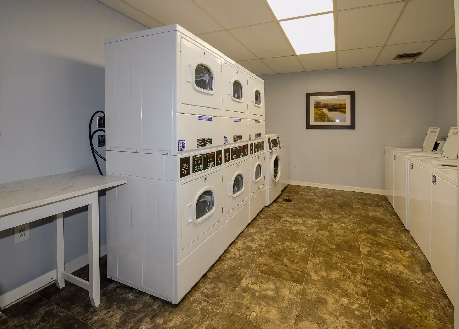 Laundry room with stacked white washers and dryers on the left side and top-loading washing machines on the right side. There is a folding table on the left wall and a framed landscape picture hanging on the back wall. The floor has a brown tile pattern and the walls are painted light gray.