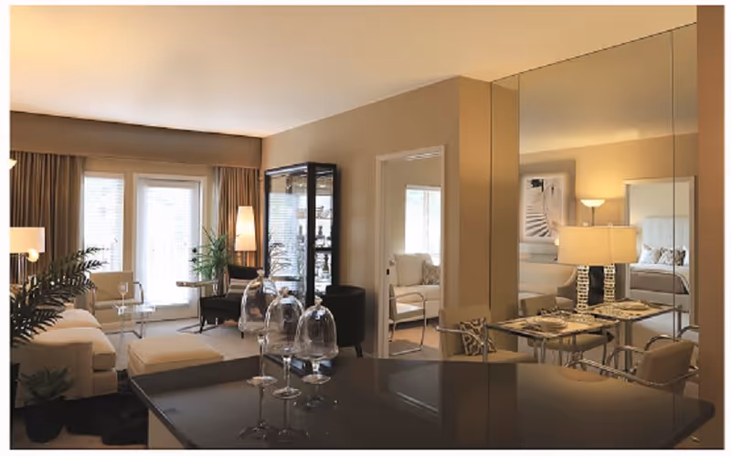 A cozy and well-lit living room area with beige walls and curtains, featuring a white sofa, a glass coffee table, a black display cabinet, and several plants. Adjacent to the living room is a dining area with a table set for two, reflected in a large mirror on the wall. A bedroom with a bed and artwork is visible through an open doorway.