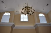 High ceiling interior with three large windows featuring arched tops and a central rectangular window. A large, ornate chandelier with multiple light bulbs hangs from the ceiling. The walls are painted beige with white trim.