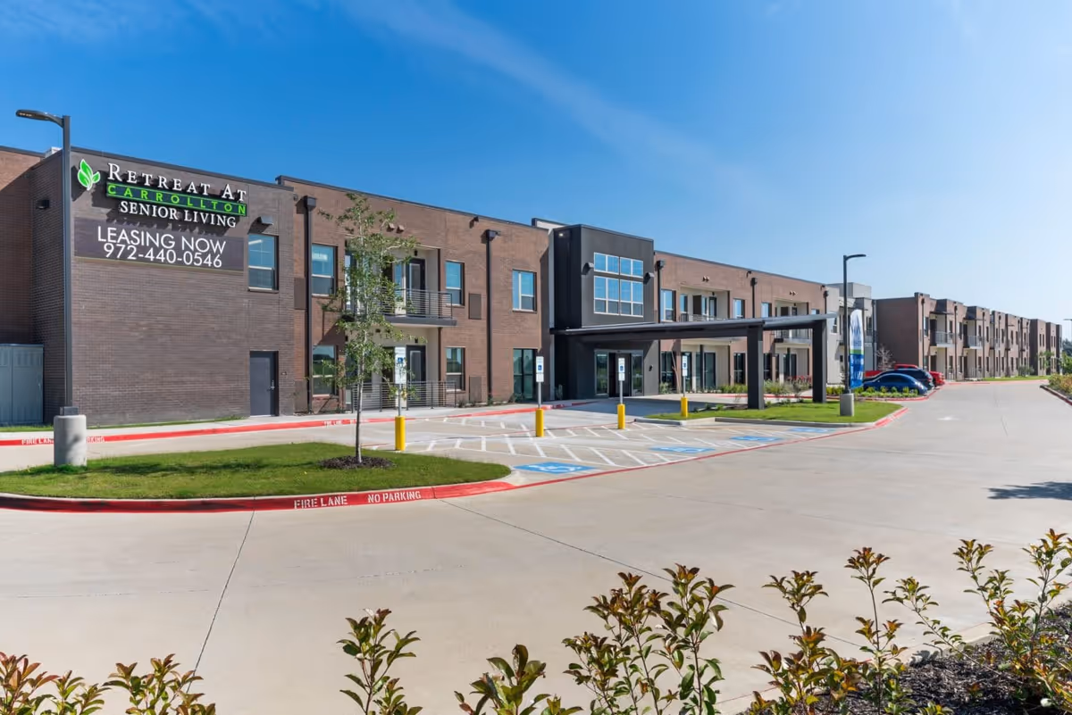 Exterior view of the Retreat at Carrollton senior living facility showing a modern two-story brick building with multiple windows and balconies, a covered entrance, parking spaces including handicapped spots, and clear blue sky.