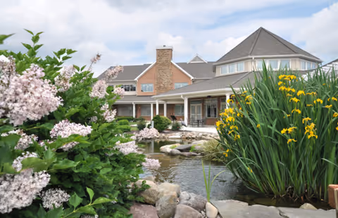 A large senior living facility building with a peaked roof and multiple windows, surrounded by a landscaped garden featuring blooming flowers, green shrubs, and a pond with rocks.