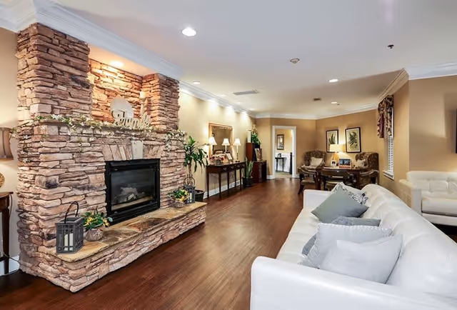 A cozy living room area featuring a large stone fireplace with decorative plants and a 'family' sign on the mantel. The room has wooden flooring, a white sofa with multiple cushions, a white armchair, and a small sitting area with two armchairs and a table in the background. The walls are painted beige with crown molding and decorated with framed artwork and plants.