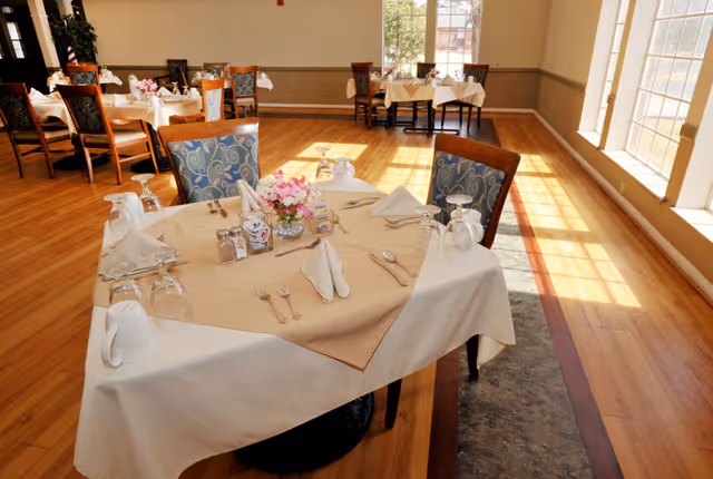 A bright dining room with wooden floors and large windows letting in natural light. Several tables are set with white tablecloths, beige overlays, folded napkins, glassware, silverware, and small floral centerpieces. Chairs with patterned upholstery surround the tables.