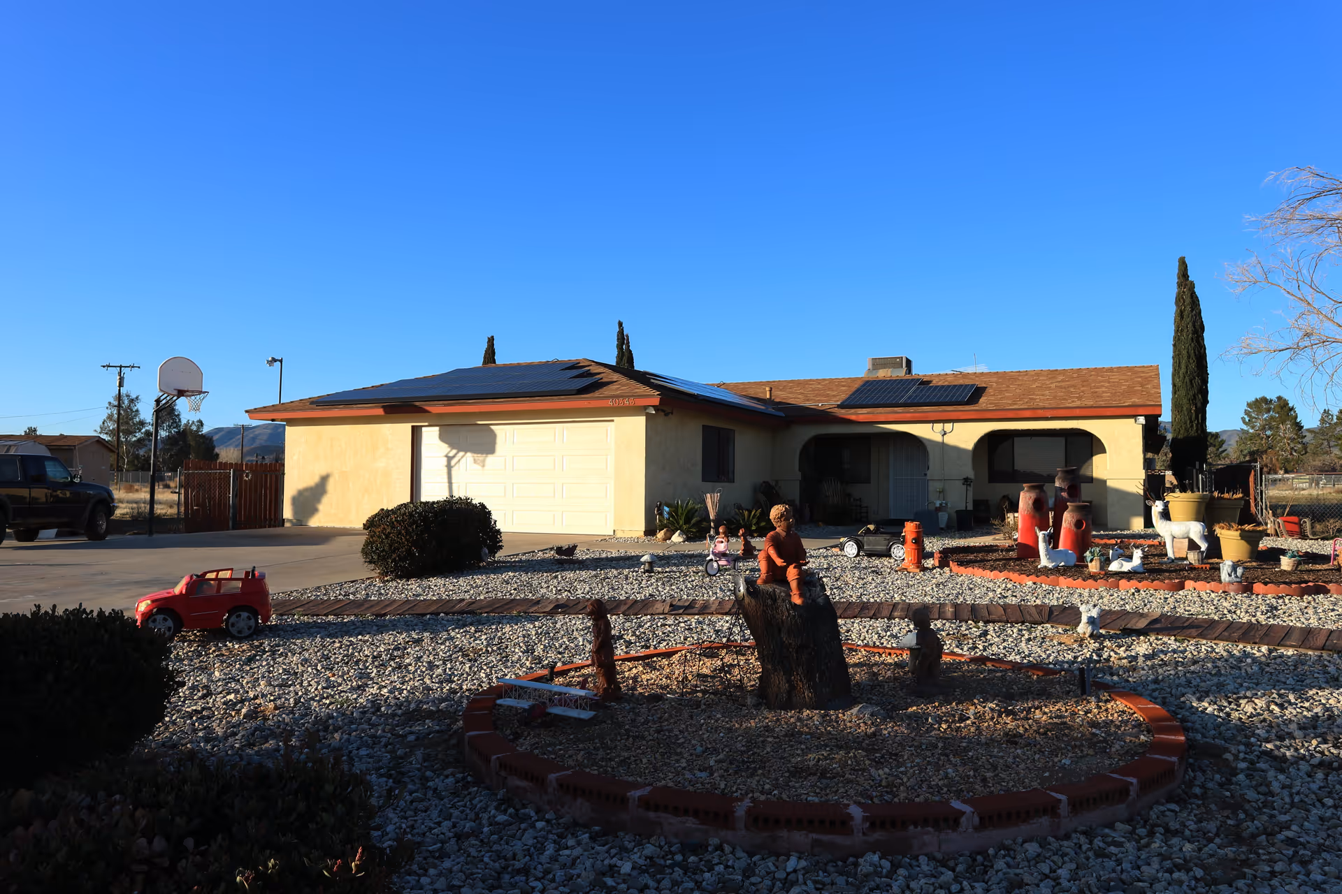 Single-story house with a beige exterior and solar panels on the roof under a clear blue sky. The front yard is covered with gravel and decorated with various garden statues, including a child sitting on a tree stump, animals, and large red vases. A basketball hoop is visible on the left side near a driveway with a black vehicle parked. A small red toy car is on the gravel near the driveway.