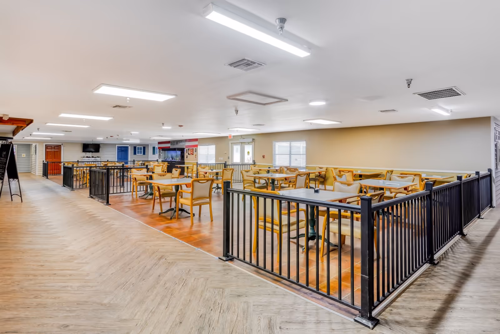 A spacious dining area in an assisted living facility with multiple tables and chairs arranged neatly. The floor is a combination of wood and tile, and the area is enclosed by black metal railings. The room is well-lit with ceiling lights, and there are windows on the far wall allowing natural light in. A television is mounted on the wall in the background.