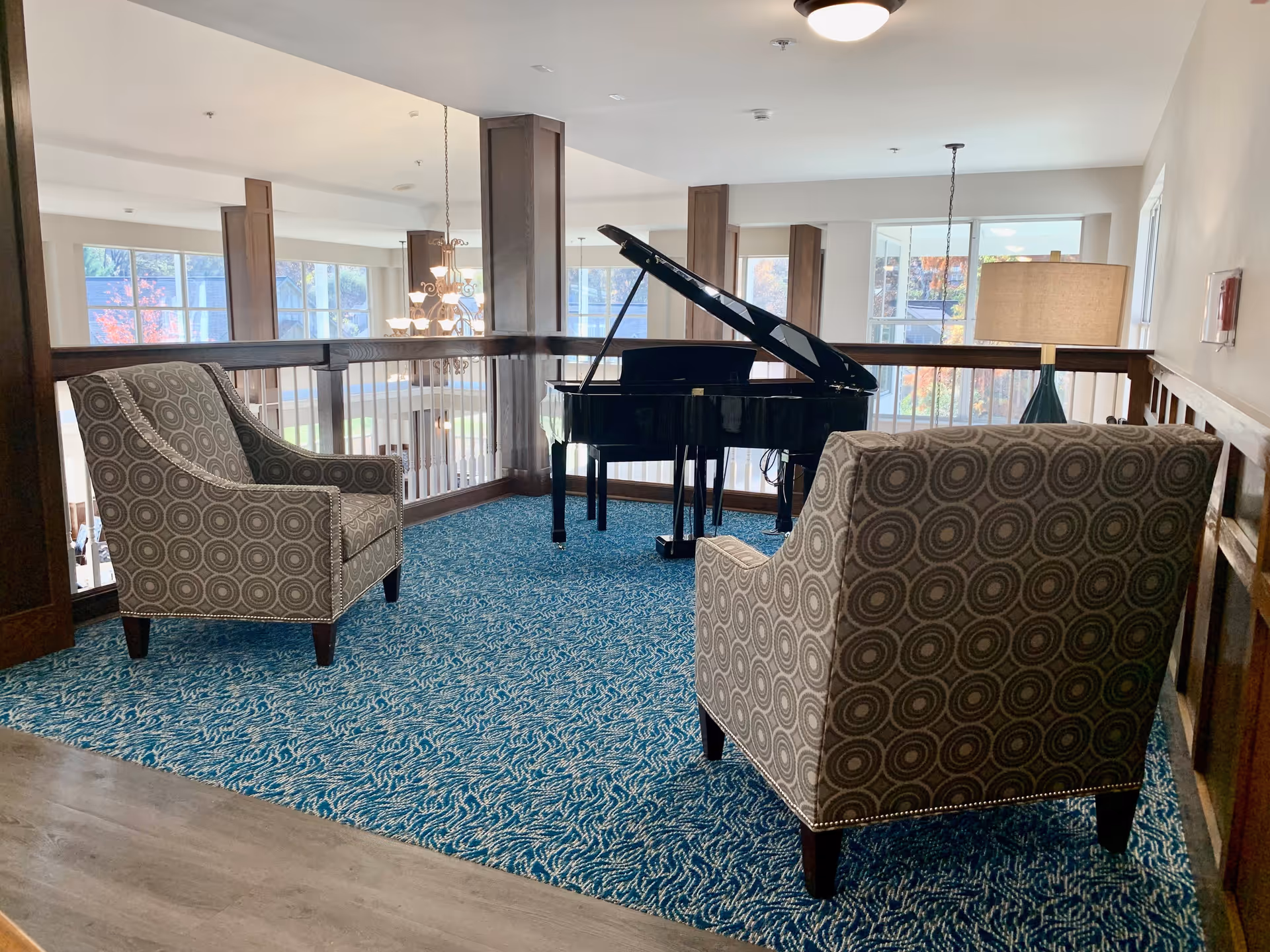 A cozy interior seating area with two patterned armchairs facing a black grand piano on a blue patterned carpet. The space is well-lit with natural light coming through large windows and a hanging chandelier visible in the background.