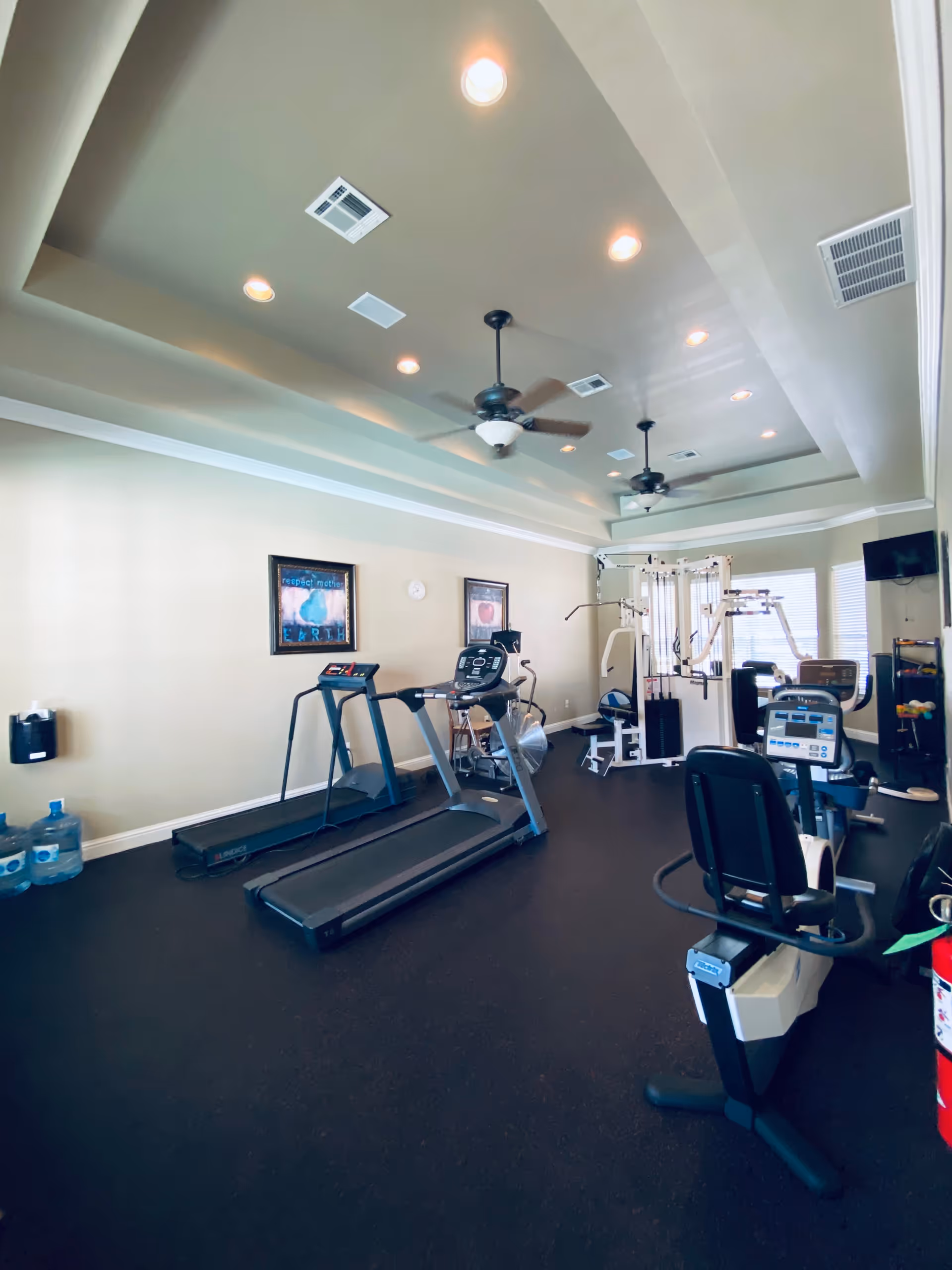 Interior view of a fitness room with exercise equipment including treadmills, stationary bikes, and a multi-function weight machine. The room has a tray ceiling with recessed lighting and ceiling fans, light-colored walls with framed artwork, and windows letting in natural light.
