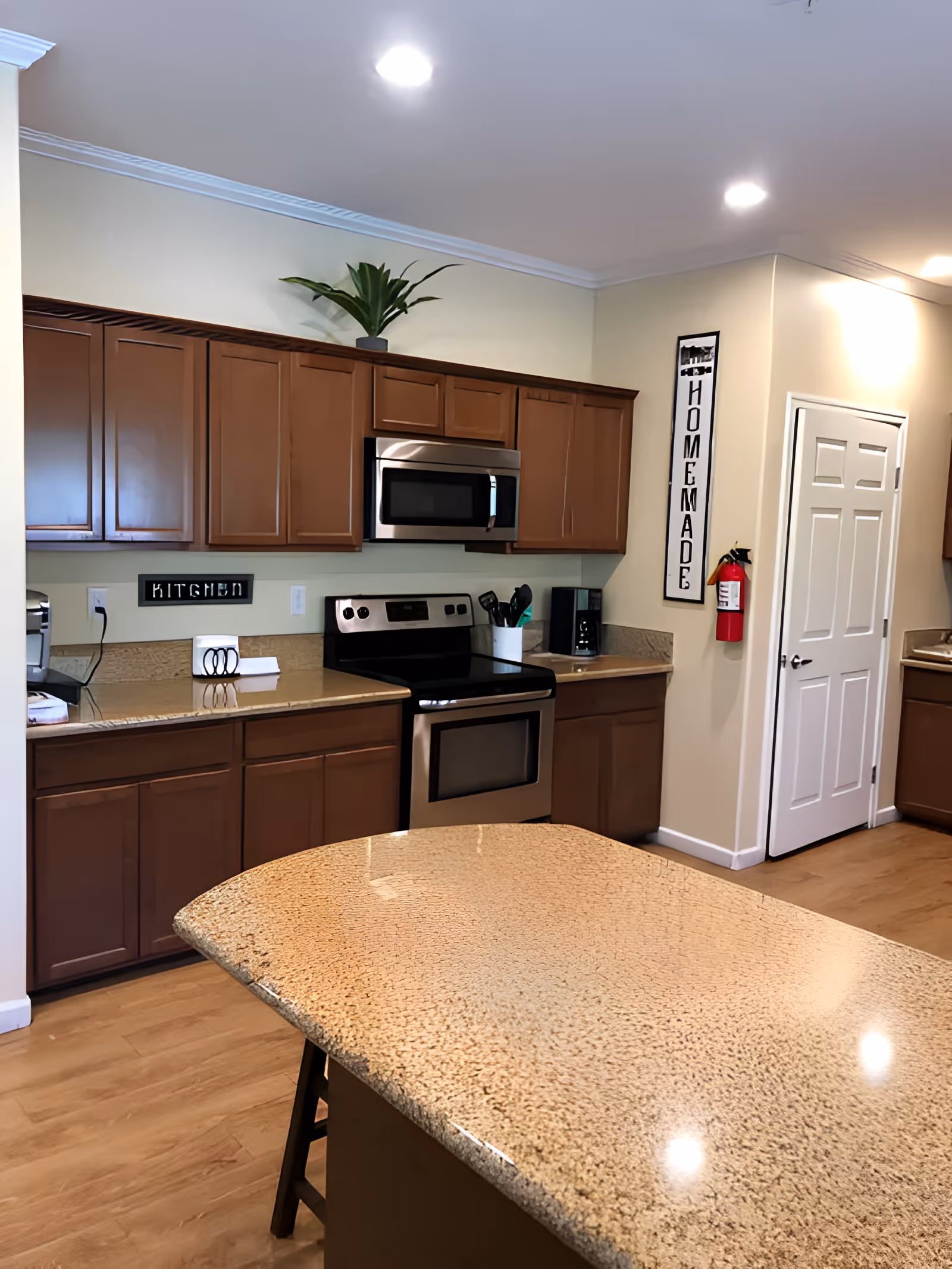 A modern kitchen with wooden cabinets, a stainless steel oven and microwave, a coffee maker, and a granite countertop island in the foreground. There is a small plant on top of the cabinets and a fire extinguisher mounted on the wall next to a door. A vertical sign on the wall reads 'HOMEMADE'.