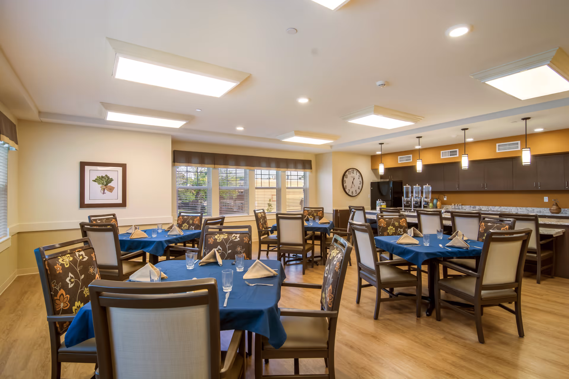 A bright dining room in a senior living facility with several tables covered in blue tablecloths, each set with folded napkins, glasses, and silverware. The room features wooden chairs with floral and beige upholstery, large windows with blinds, a wall clock, and a kitchen area with dark cabinets and pendant lighting.