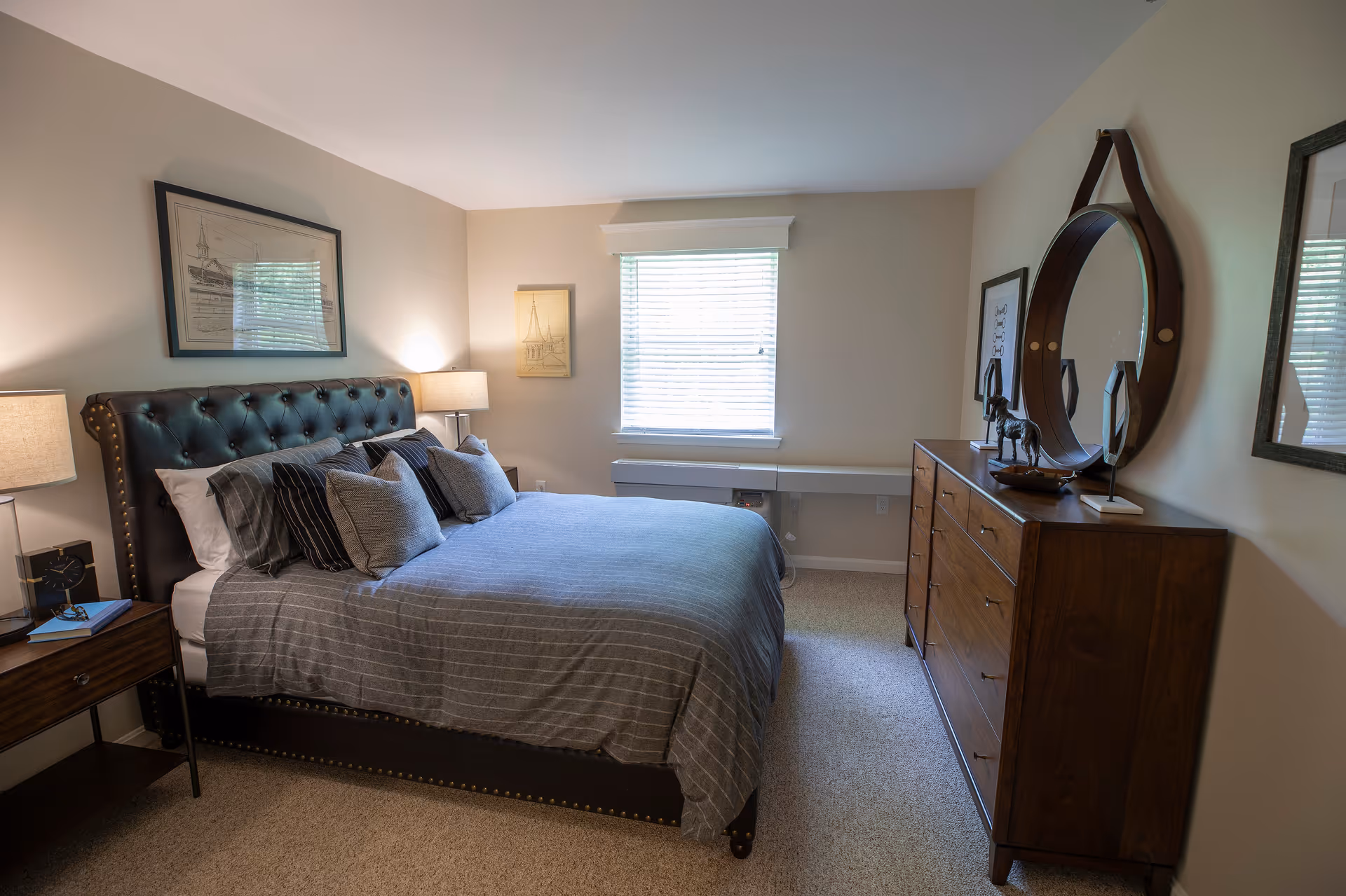 A cozy bedroom with a large bed featuring a tufted dark leather headboard and multiple pillows. There are two bedside tables with lamps, framed artwork on the walls, a window with blinds, and a wooden dresser with a round mirror and decorative items on top.