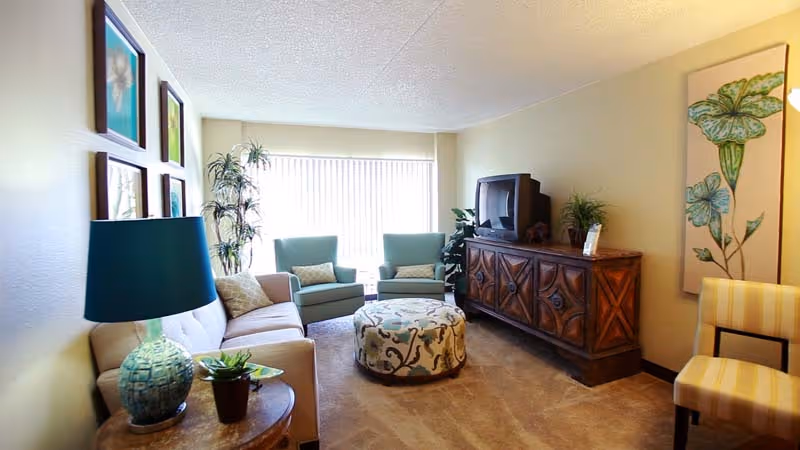 A cozy living room with a beige sofa, two green armchairs, a round patterned ottoman, a wooden side table with a blue lamp and a small plant, a wooden cabinet with a TV on top, and wall art featuring floral designs. The room is softly lit with natural light coming through vertical blinds on a large window.