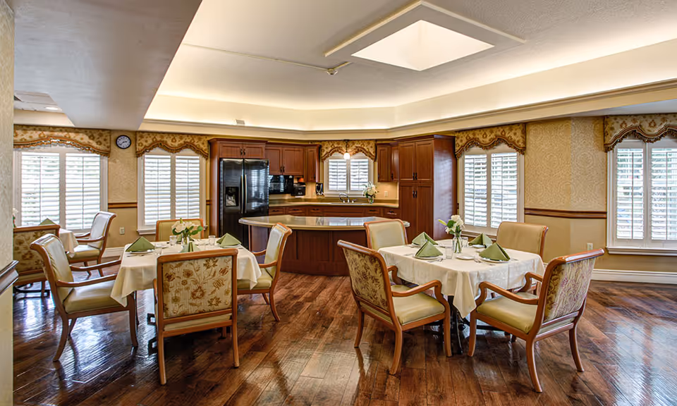 A bright dining area with two tables set with white tablecloths, green napkins, and small flower vases. The room features wooden chairs with floral upholstery, large windows with white shutters, and a kitchen area with wooden cabinets and a black refrigerator in the background. The floor is polished wood and the ceiling has recessed lighting.