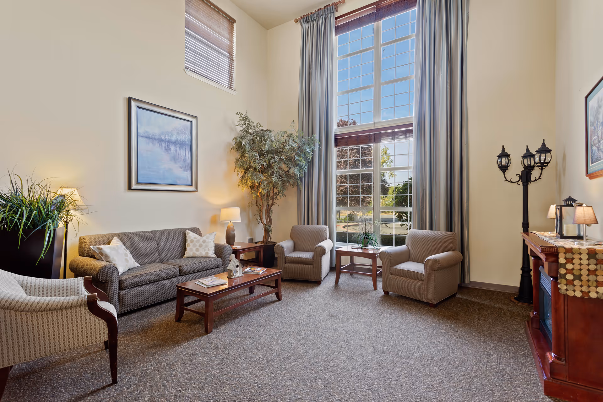 A cozy senior living facility common area with a sofa, two armchairs, a coffee table, a side table with a lamp, a tall indoor plant, large windows with curtains letting in natural light, a framed painting on the wall, and a decorative lamp post-style floor lamp next to a wooden cabinet with a table runner and small lamps.