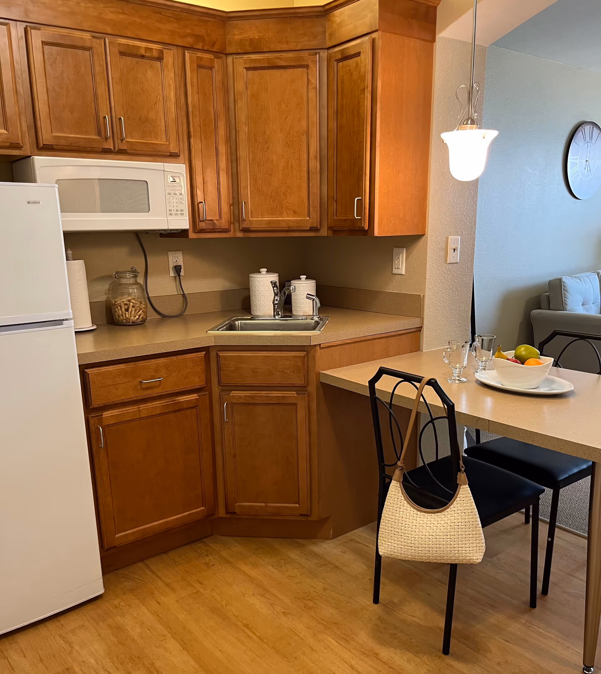 A compact kitchen area with wooden cabinets, a white refrigerator, a white microwave, and a small sink. There is a countertop extending to form a small dining area with two black chairs, one of which has a beige woven handbag hanging on it. On the countertop, there is a white bowl with fruit and two clear glasses. A hanging light fixture is visible above the dining area, and part of a living room with a clock on the wall and a gray couch can be seen in the background.