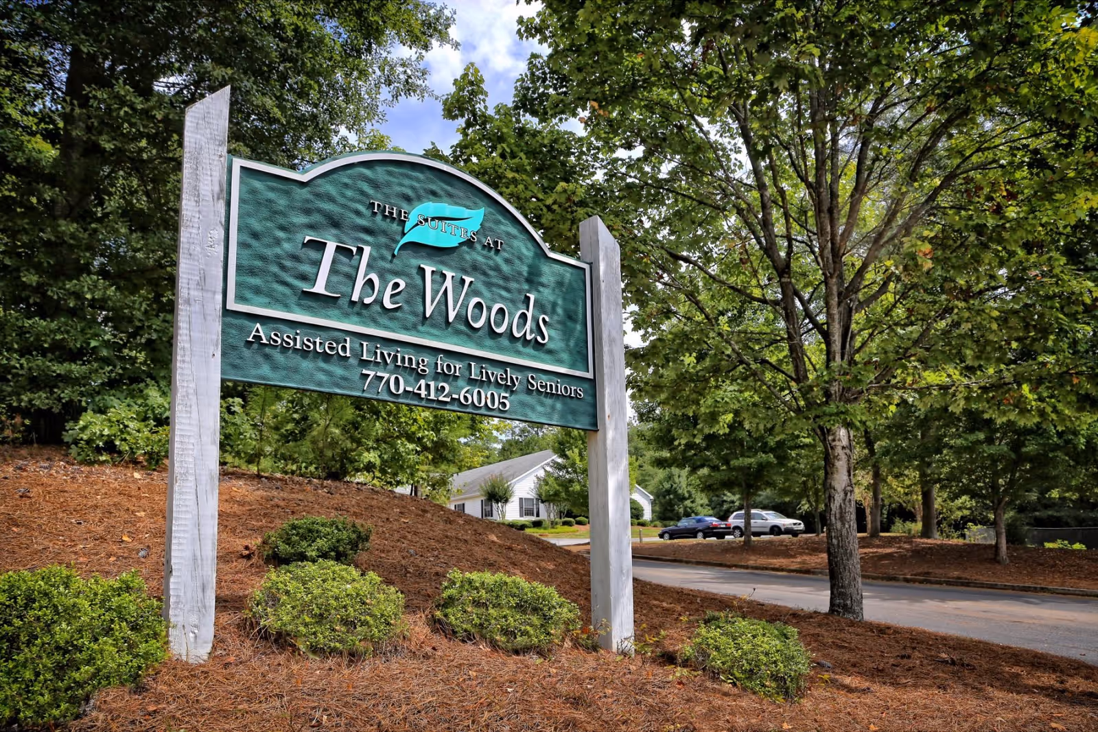 A green sign for The Woods Assisted Living for Lively Seniors with a phone number, surrounded by trees and bushes near a driveway with parked cars.