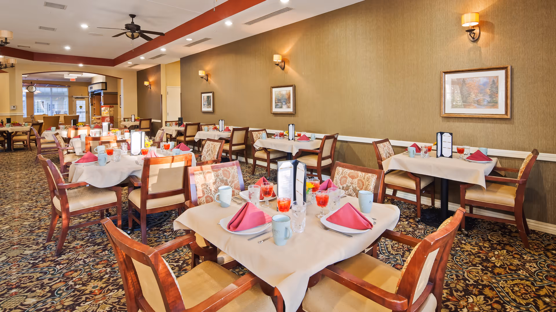 A dining room in a senior living facility with multiple tables set for meals. Each table has beige tablecloths, pink folded napkins, glasses with red beverages, mugs, and menus. The room features patterned carpet, beige walls with framed artwork, and ceiling fans with lights. The space is well-lit with natural light coming from windows in the background.