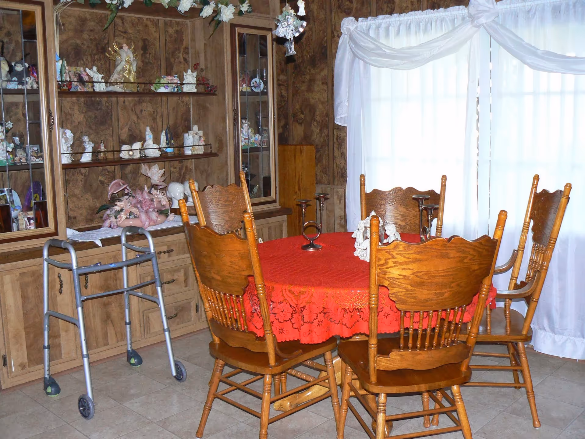 A dining room with a round wooden table covered with a red lace tablecloth surrounded by six wooden chairs. Behind the table is a wooden cabinet with glass shelves displaying various figurines and decorations. A walker is positioned near the cabinet. White curtains cover a large window in the background.