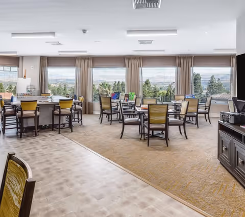 A bright and spacious common area with multiple tables and chairs arranged for group seating. Large windows with blinds and curtains allow natural light to fill the room and offer a view of trees and distant hills. The room has a mix of carpet and tiled flooring, and there is a TV on a stand to the right side of the image.