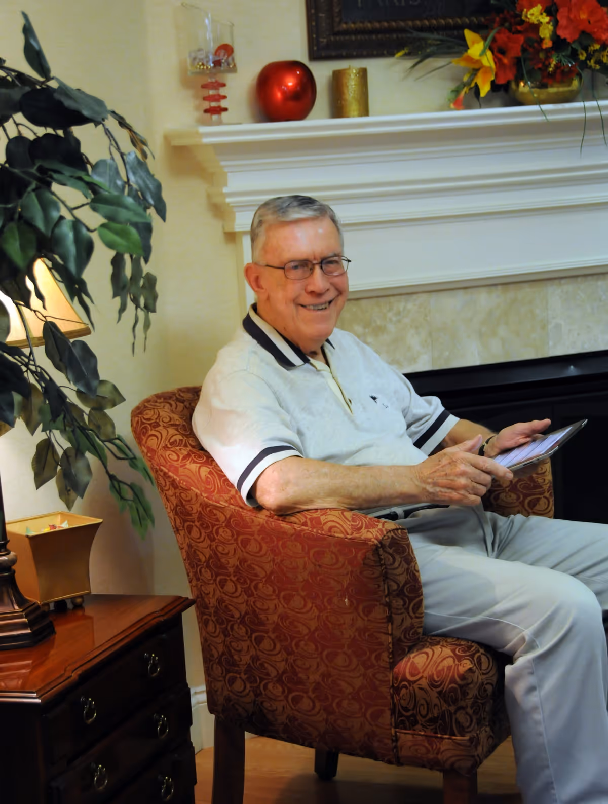 An elderly man with glasses sitting in a patterned red armchair in a cozy living room. He is smiling and holding a tablet. Behind him is a white fireplace mantel decorated with a red apple ornament, a gold candle, and a floral arrangement. A leafy green plant and a table lamp are visible to the left.