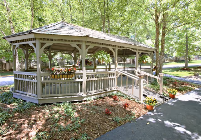 A wooden gazebo with a shingled roof situated in a garden area surrounded by trees. The gazebo has railings and a ramp for accessibility, with hanging plants and flower pots decorating the structure. A paved pathway runs alongside the gazebo.