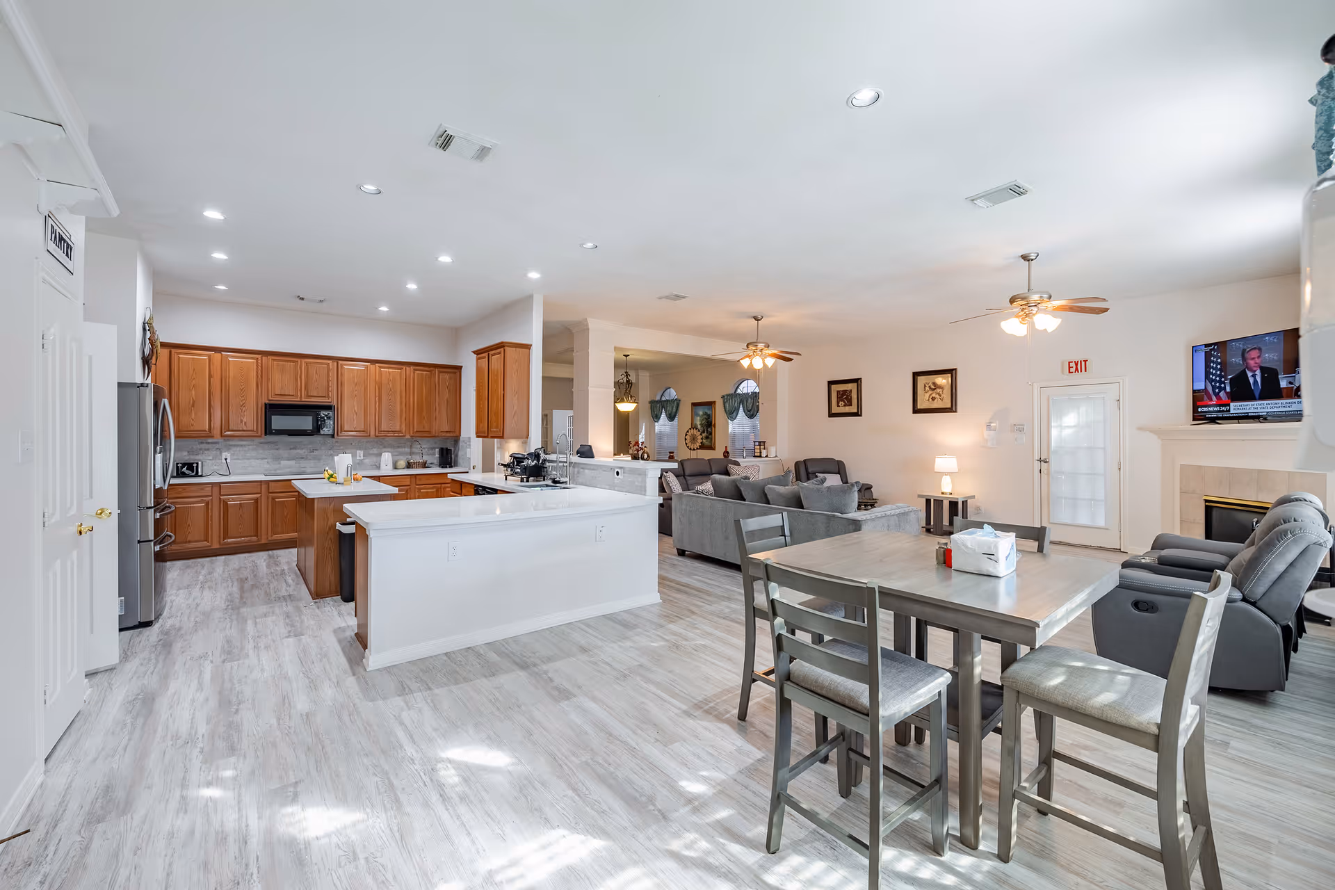 Open-plan interior showing a kitchen with wooden cabinets and an island, a dining table, and a living area with sofas and a TV.