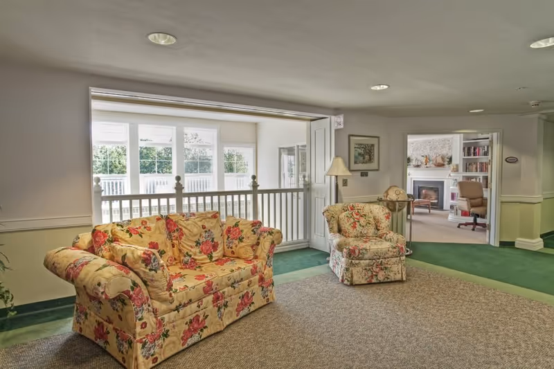 Sunlit lounge with floral-pattern sofa and matching armchair, carpeted floor, large windows and a doorway leading to a fireplace room.