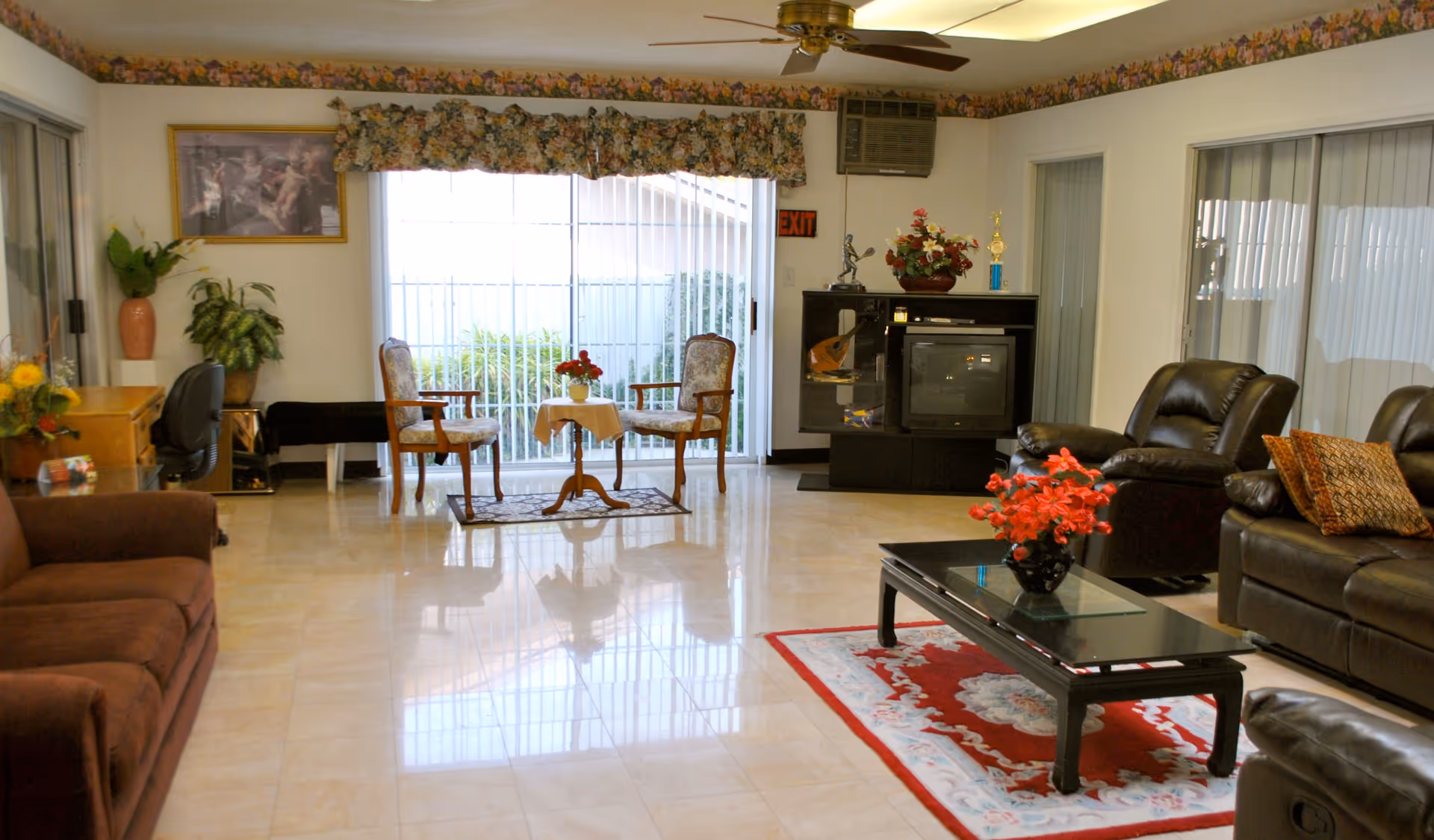 A spacious living room in a senior care facility with a brown sofa on the left, two armchairs with a small table and flowers near a large window with vertical blinds, a black entertainment center with a TV and decorative items, and a black coffee table with a red flower arrangement on a red and white rug. The room has tiled floors, a ceiling fan, and floral wallpaper border near the ceiling.