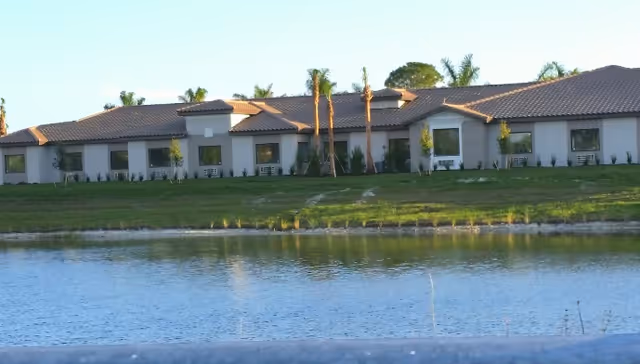 Single-story stucco building with a tiled roof and palm trees along a grassy shore reflected in a pond.