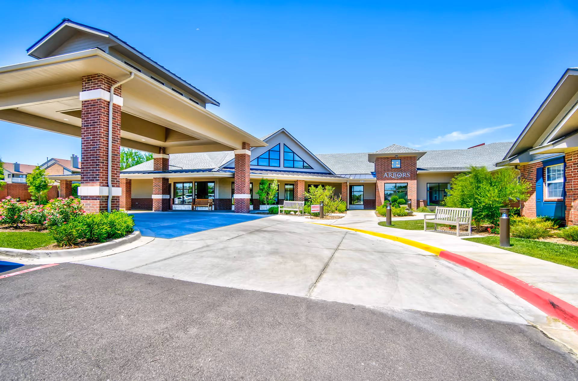 Front exterior view of a senior living facility named Arbors with a covered entrance supported by brick columns, surrounded by landscaped greenery and benches under a clear blue sky.
