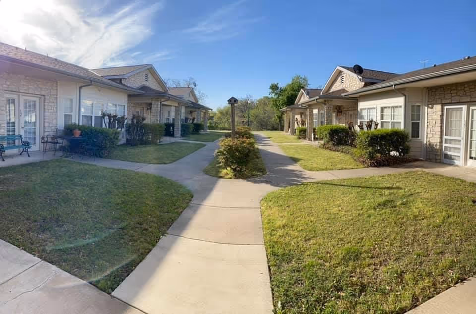 Outdoor view of a senior living facility courtyard with paved walkways, green lawns, shrubs, and single-story stone buildings with porches and windows under a partly cloudy sky.