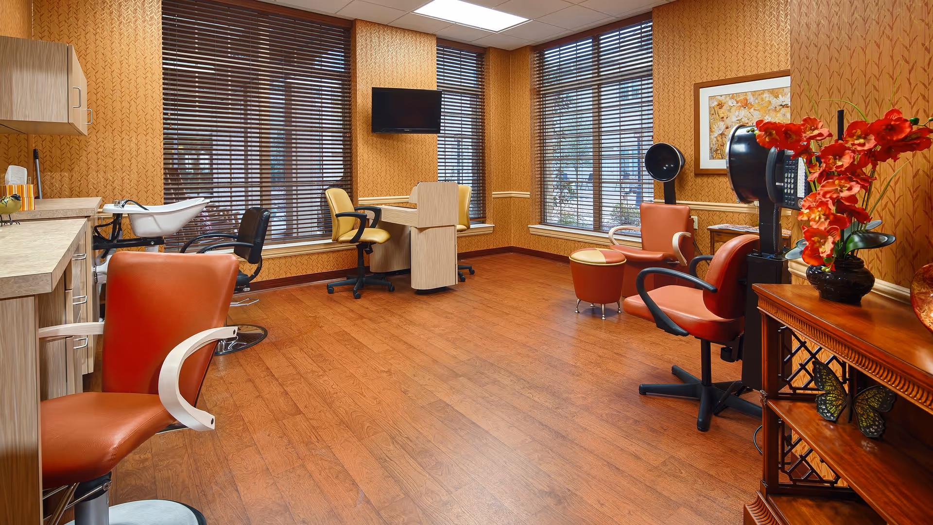 Interior view of a salon area in a senior living facility with wooden flooring, orange salon chairs, a hair washing station, a TV mounted on the wall, large windows with blinds, and a wooden cabinet with red flowers and decorative items.