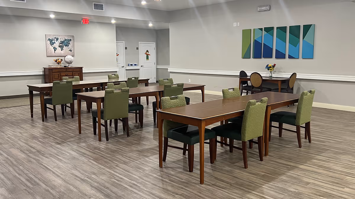 A spacious dining room with several wooden tables and green upholstered chairs arranged neatly on a wood-patterned floor. The walls are light gray with white trim, decorated with a world map artwork and a set of abstract blue and green panels. There is a small table with flowers and two chairs in the corner, and two closed white doors in the background.