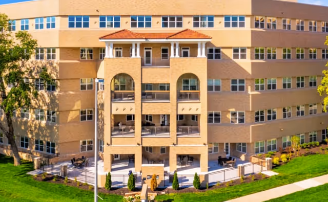 Exterior view of a multi-story senior living facility building with beige brick walls, multiple windows, and balconies. The building has a small outdoor patio area with tables and chairs, surrounded by green grass and landscaping.