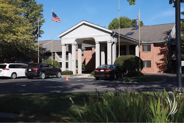 Exterior view of a brick building with a white covered entrance supported by columns. Several cars are parked in front, and there are flagpoles with flags near the entrance. Trees and greenery surround the area under a clear blue sky.