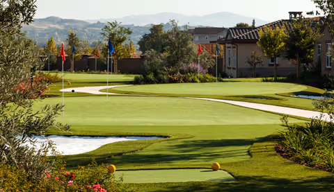 Well-maintained putting green with several flagsticks, a sand bunker, walking paths, landscaping and nearby residential buildings with hills in the background.
