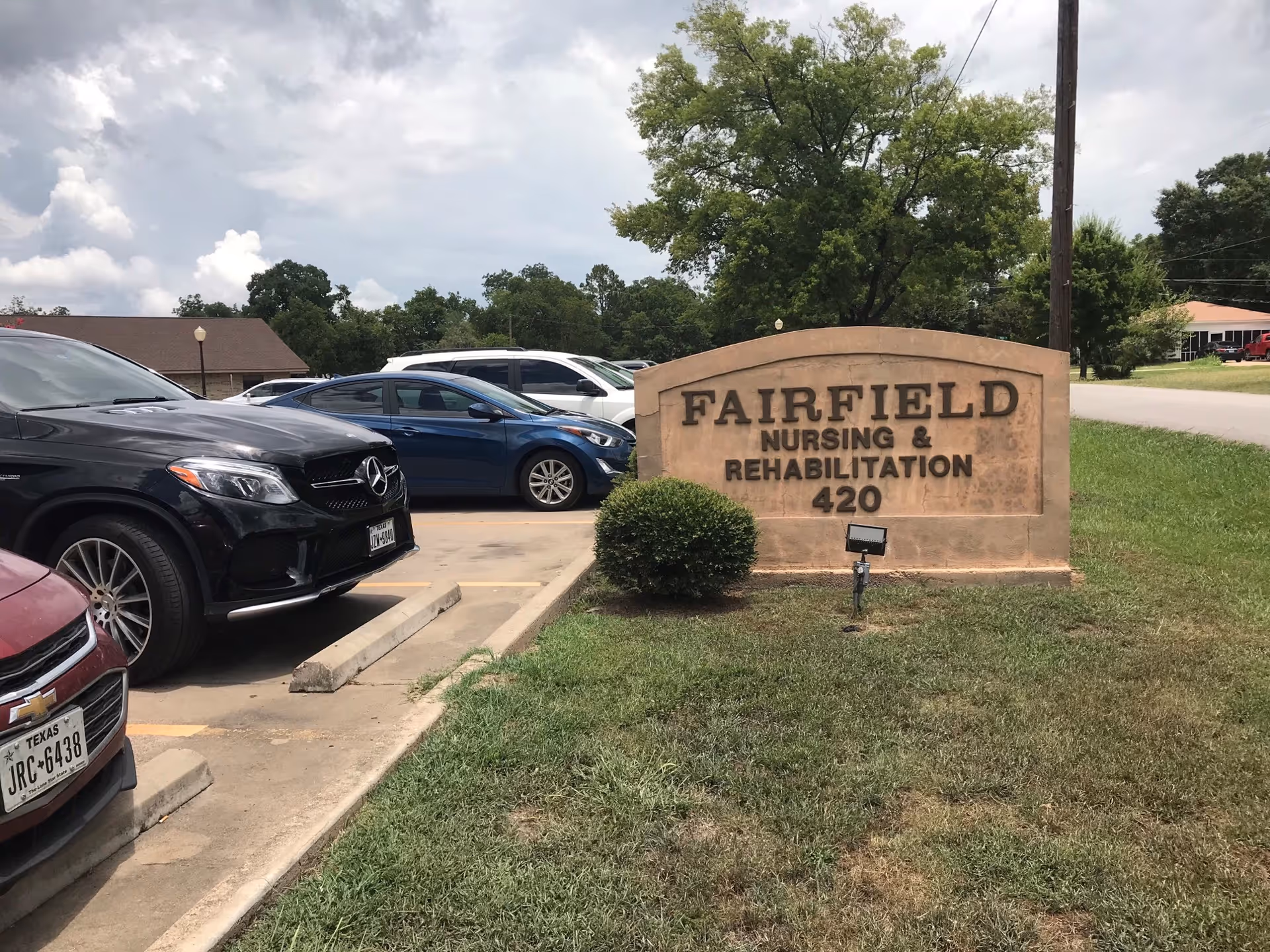 Stone sign reading 'FAIRFIELD NURSING & REHABILITATION 420' next to a parking lot with parked cars and a grassy lawn.