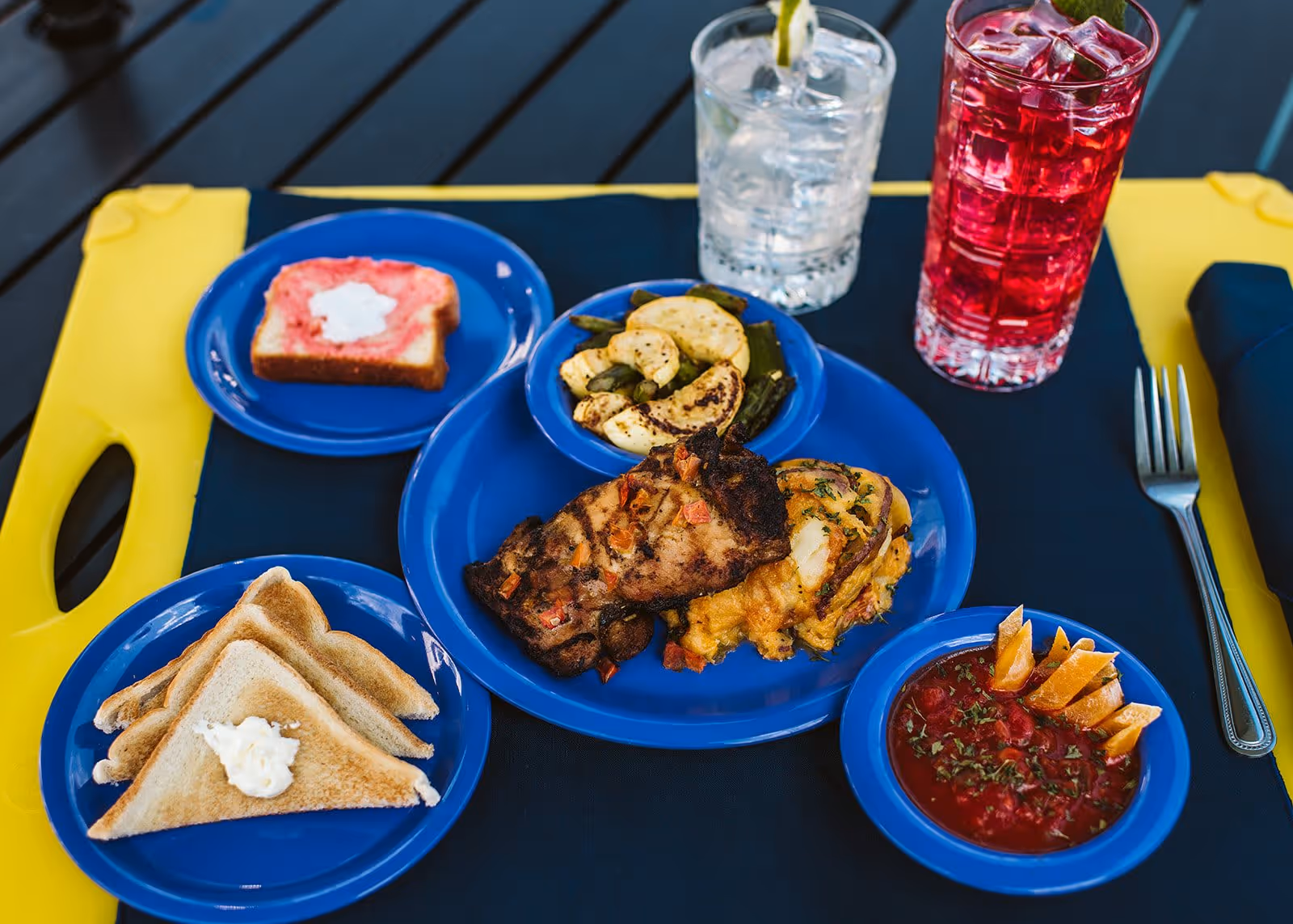 A meal served on blue plates on a dark blue placemat with a yellow tray underneath. The meal includes grilled meat with a side of roasted vegetables and a cheesy potato dish, a small bowl of tomato-based soup garnished with herbs and carrot sticks, toasted bread with a dollop of butter, a slice of bread with pink spread and white topping, a glass of water with ice and a lemon wedge, and a glass of red beverage with ice. A fork and a folded dark blue napkin are placed to the right.