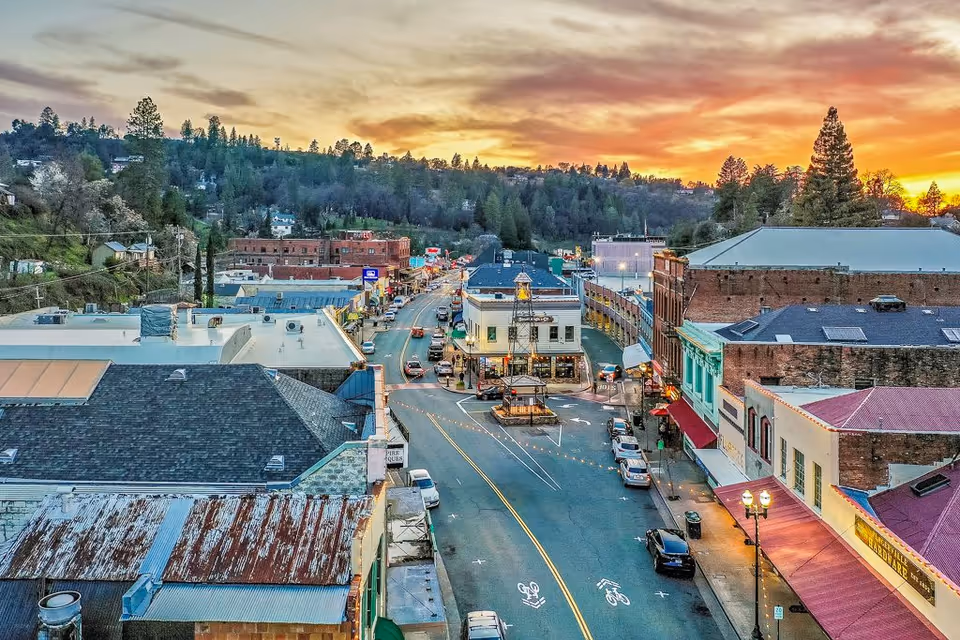 Aerial view of a small downtown main street with brick buildings, parked cars, and a colorful sunset sky.