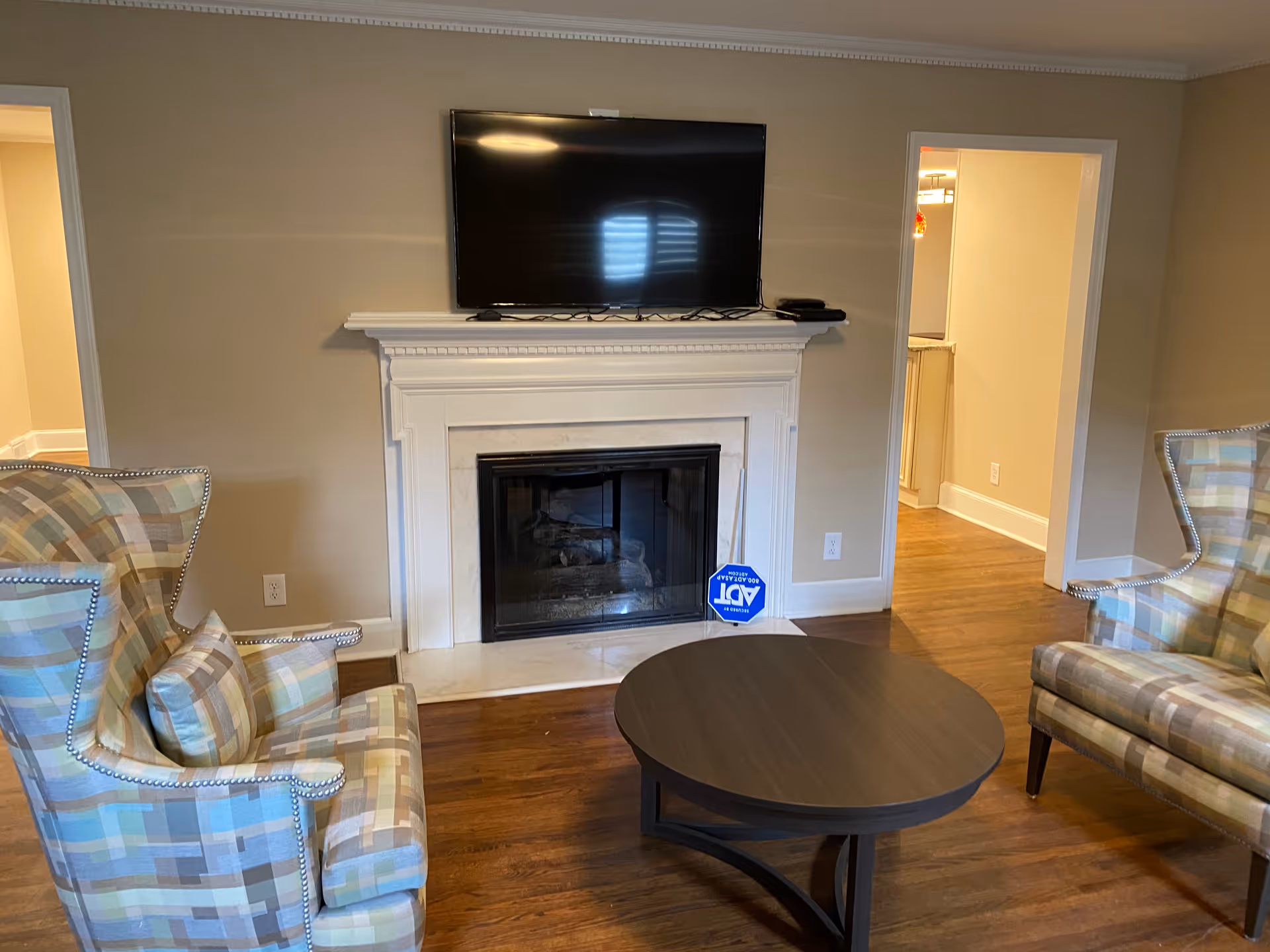 Living room with a fireplace and wall-mounted TV, two patterned armchairs and a round coffee table on hardwood floors.