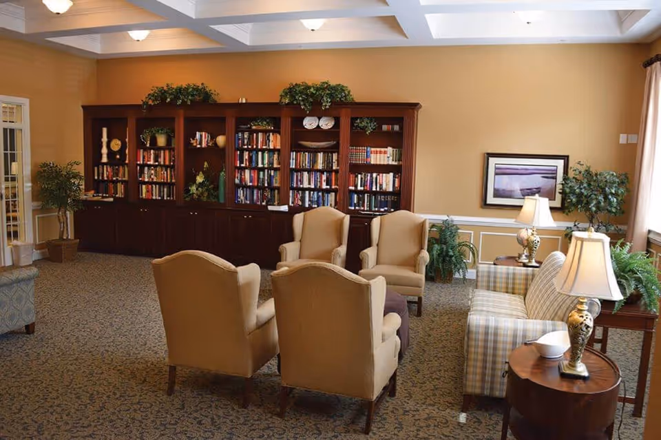 A cozy living room area with four beige armchairs arranged around a small ottoman, a plaid sofa, two side tables with decorative lamps, and a large wooden bookshelf filled with books and decorative items against a beige wall. There are also several potted plants and a framed picture on the wall.