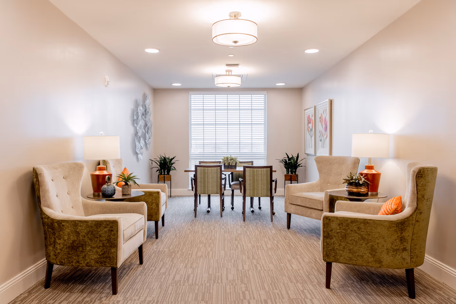 A well-lit senior living common area with four beige armchairs arranged in pairs facing each other, each pair separated by a small round table with decorative plants and lamps. At the far end, there is a window with blinds and a table with four chairs. The walls are decorated with framed floral artwork and a metal wall sculpture.